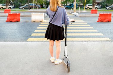 Scooter 'ı olan genç bir kızın yaya geçidinde yeşil ışık beklerken görüntüsü. Şehir caddesinde insanlar elektrikli scooter kullanıyor..
