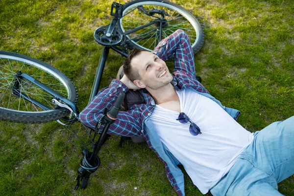 stock image Young man lying on lawn next to bike in summer park. happy person resting after cycling, eyes closed, enjoying silence in nature. Sport, outdoor activity, fitness concept