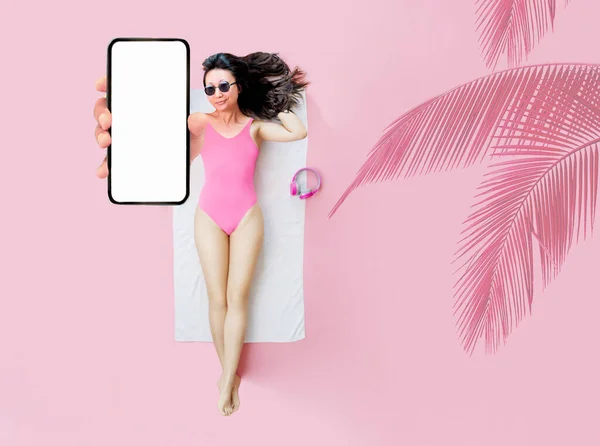 身穿粉色泳衣的全尺寸亚裔顶级女性躺在浴巾酒店游泳池上 展示了用纯粉色背景隔离的空智能手机屏幕 暑假海上休息日光浴的概念 — 图库照片