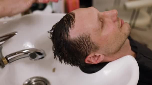 美容室で美容師に洗髪される若い男性のクライアント チューブを通して撃たれた 専門的なサービスと髪の洗浄とマッサージ 理髪店だ 高級美容室に設置された洗面器 — ストック動画