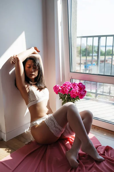 白い壁の近くに花でポーズをとるランジェリー姿のアジア系の若い女性 太陽の光と色 — ストック写真