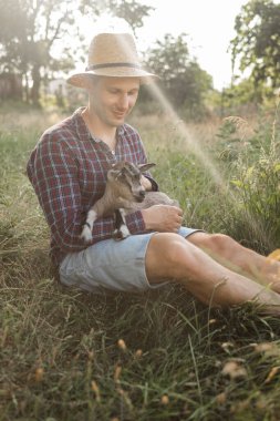 Kafkas görünümlü mutlu bir adam, bir çiftçi en sevdiği keçisini kucaklar ve kucaklar. Küçük bir keçi. Yazın kırsal alan kavramı. Açık hava eğlencesine yakın.
