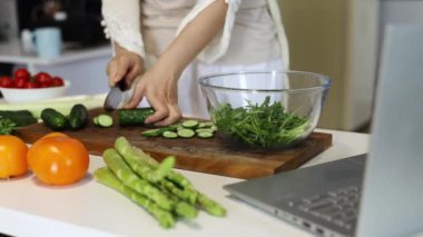 Modern, temiz bir ev mutfağında hazırlanmış taze sebzelerle birlikte salatalık kesen bir kadın. 