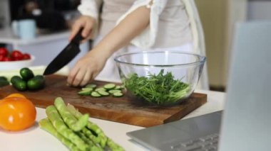 Kadın taze salata pişiriyor. Salatalık kesiyor. Yemek tarifini bilgisayarda izliyor.