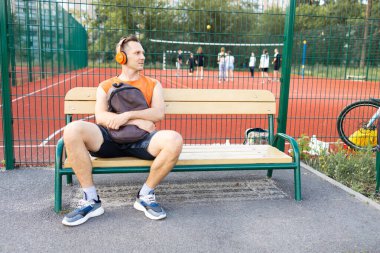 Turuncu tişörtlü yakışıklı bir adam parkta dinleniyor ve kulaklıkla müzik dinliyor.