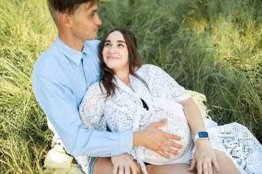 Happy pregnant couple sitting on the grass in park. Happy and young pregnant couple hugging in nature.
