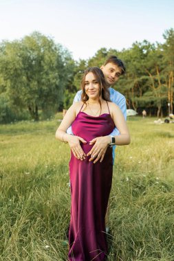 Happy pregnant couple sitting on the grass in park. Happy and young pregnant couple hugging in nature.