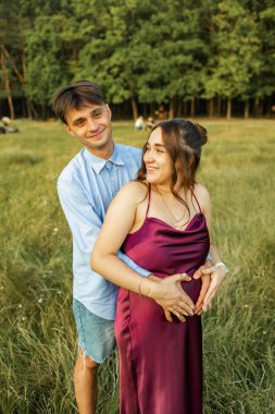 Happy pregnant couple sitting on the grass in park. Happy and young pregnant couple hugging in nature.