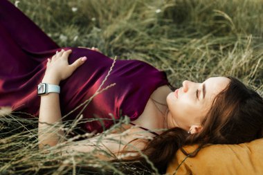 Happy pregnant woman lying on grass in summer park outdoors.