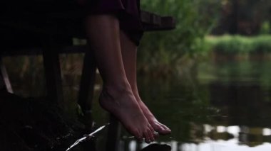 Bir kadın gölün kenarında dinleniyor, tahta bir iskelede oturuyor, bacaklarını suyun yüzeyinde sallıyor..