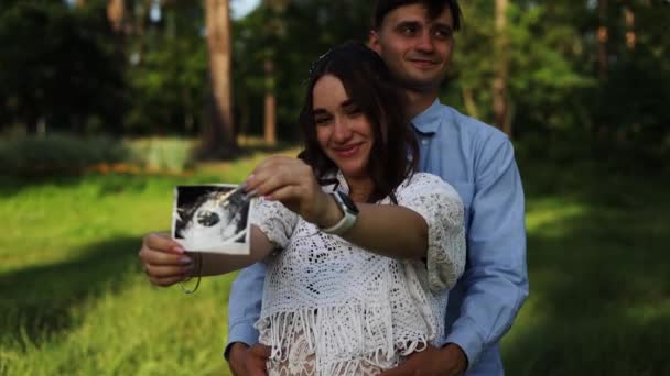 快乐的怀孕夫妇坐在公园的草地上 年轻孕妇在大肚子里拿着未出生婴儿的超声照片 — 图库视频影像