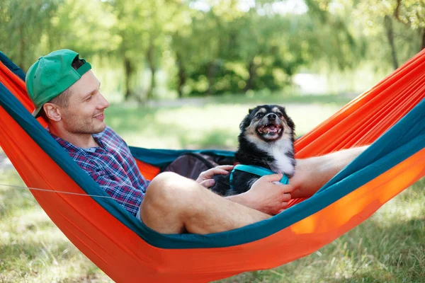 Man with dog resting in hammock near lake in summer day.