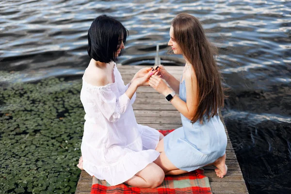 川岸の桟橋に座っている2人のティーンエイジャーの女の子は 夏に楽しい時間を過ごしています ハッピーガールフレンド 湖の近くのアウトドアをリラックス — ストック写真