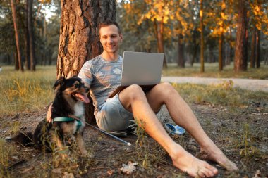 Parkta dizüstü bilgisayarla çalışan köpeği olan genç bir adam..