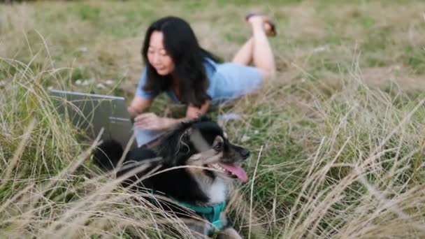 草の上に横たわっている小さな犬を切り アジアの女の子は彼の隣で働いています — ストック動画