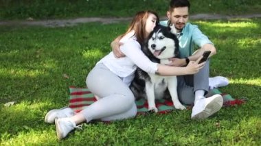 Şirin köpekli genç çift yaz parkında yeşil çimlerde selfie çekiyor..