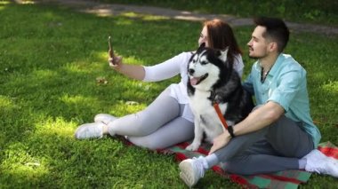 Şirin köpekli genç çift yaz parkında yeşil çimlerde selfie çekiyor..