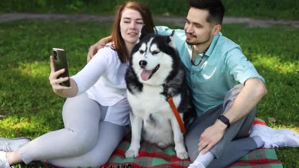 一对年轻夫妇带着可爱的哈士奇狗在夏日公园的绿草上自拍 — 图库视频影像
