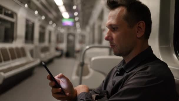 一位严肃的高加索男性公司员工在下班后深夜驾驶着一辆移动中的现代城市地铁列车 在手机上阅读新闻 — 图库视频影像