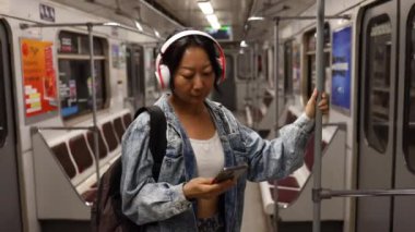 Genç Asyalı kız metroya biniyor telefona bakıyor ve kulaklıkla müzik dinliyor. Metroda akıllı telefon kullan.. 