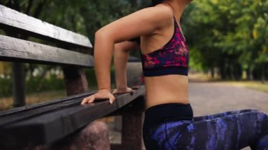 Orta yaşlı Asyalı kadın şehir parkında bank dalışı egzersizleri yapıyor. Bayan sporcu triseps kaslarını dışarıda çalıştırıyor. Uzun menzilli çekimler. Vücut ağırlığı eğitimi dışarıda.