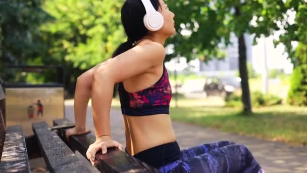 中世のアジアの女性は 市の公共公園でベンチディップ演習を行っています 女性アスリートのトレーニングは 筋肉を屋外でトリプルします 全長撮影をハンドヘルドする 外でのボディウェイトトレーニング — ストック動画