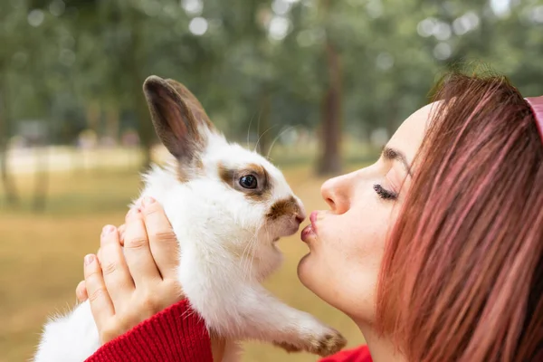 一位穿着红色毛衣的年轻女子吻了公园里的一只白兔 — 图库照片