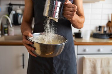 Evdeki kaseye un karıştıran bir adamın yakın çekimi. Önlüklü genç adam mutfakta elmalı turta pişiriyor, fırında un ekliyor..