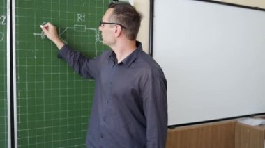 Beyaz adam öğretmen karatahtaya geometri denklemi yazıyor, öğrencilerle konuşuyor.