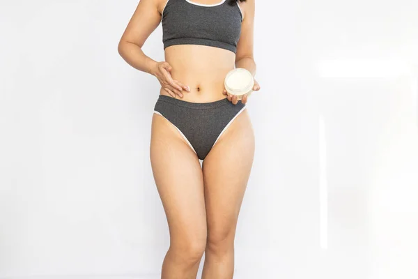 Stretch marks and cellulite on body. woman in panties applying moisturising body cream on her legs, using anti-cellulite lotion or sunscreen while standing on white background, cropped.