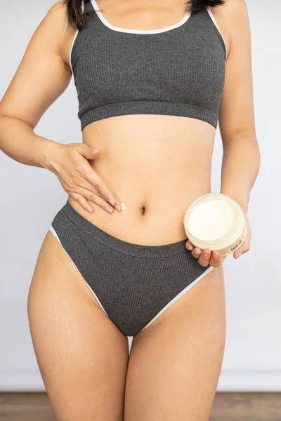 Stretch marks and cellulite on body. woman in panties applying moisturising body cream on her legs, using anti-cellulite lotion or sunscreen while standing on white background, cropped.