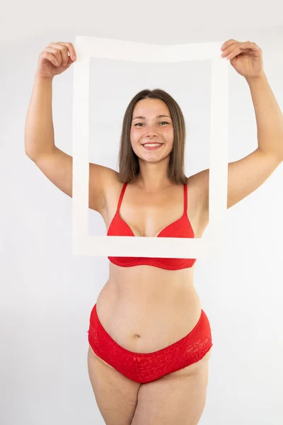 Body positivity and self love concept. Waist up shot of joyful body positive young woman holding frame feels confident love own body wears red lingerie poses over white wall