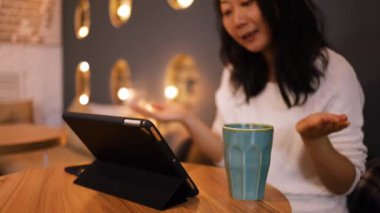 Gülümseyen Asyalı kız dizüstü bilgisayarla çevrimiçi video görüşmesi sırasında konuşuyor ve el sallıyor, kahve dükkanında oturuyor. Neşeli iş kadını taşınabilir not defteriyle internette konferans veriyor..