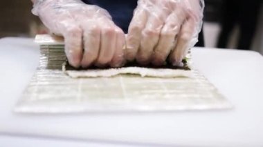 Şef restoranın mutfağında Japon suşisi yuvarlıyor. Şef, Japon restoranında suşi yapmak için hasırı kullanıyor. Şef Japon suşi rulosuna salatalık koyuyor.