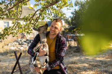 Kafkas görünümlü mutlu bir adam, bir çiftçi en sevdiği keçisini kucaklar ve kucaklar. Küçük bir keçi. Yazın kırsal alan kavramı. Açık hava eğlencesine yakın.