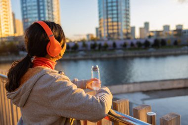 Genç, güzel ve mutlu bir Asyalı kadın spor kıyafetleri içinde kulaklıkla müzik dinliyor ve şehir köprüsünde egzersiz yaptıktan sonra su şişesi tutuyor sağlıklı yaşam tarzı konseptiyle.