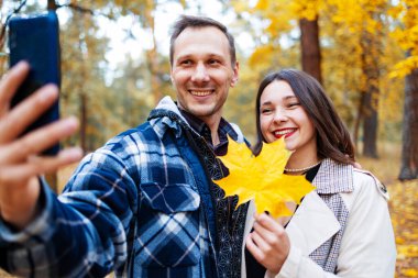 Çekici genç çift birlikte zaman geçirip sonbahar parkında akıllı telefonlardan selfie çekiyor..  