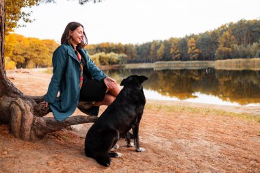 Kız ve evcil hayvanı göl kenarında yürüyüş yapıyor. Sonbahar ormanında kütüğün üzerinde büyük siyah köpeğiyle oturan mavi ceketli kız..