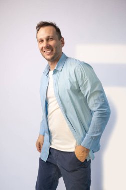 Mavi gömlekli yakışıklı beyaz arka planda kameraya gülümsüyor.