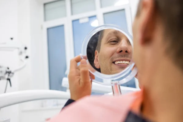 Modern klinikte diş tedavisi gören yakışıklı beyaz adam Stomatoloji Kabinesinde oturan erkek hasta ve yeni gülümsemesinin tadını çıkarıyor.