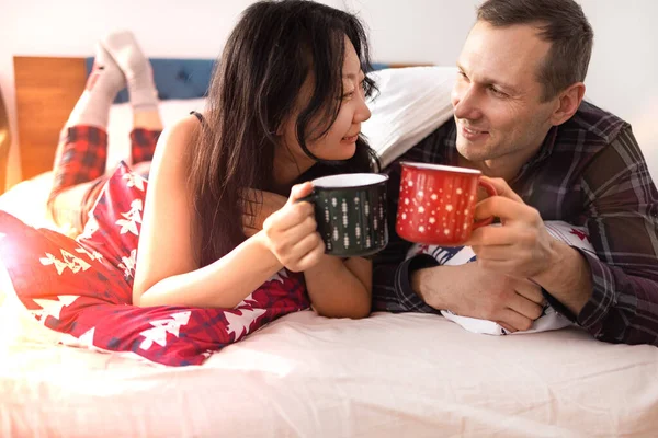 Couple in love lying in bed and drinking coffee with Christmas cups. Man and woman in new year's morning.
