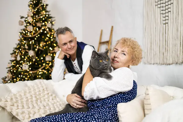 Happy senior couple with cat sitting on sofa at home near christmas tree