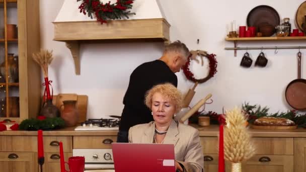 上了年纪的50多岁的妻子用手提电脑从家里的办公室远程工作或远程学习 而她的大丈夫则在厨房做早餐 帮助料理家务 — 图库视频影像