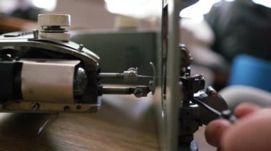 Tamirci ustası atölyedeki dikiş makinesini test ediyor. Masada oturuyor, yan görüyor. Adam dikiş makinesinin içine bakıyor, tamir etmeye çalışıyor, elleri yakın..