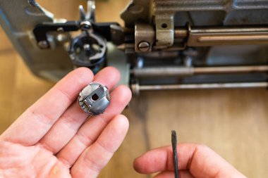 Tamirci ustası atölyedeki dikiş makinesini test ediyor. Masada oturuyor, yan görüyor. Adam dikiş makinesinin içine bakıyor, tamir etmeye çalışıyor, elleri yakın..