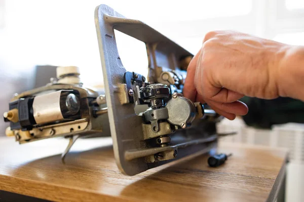 Repairman master is testing disassembles sewing machine in workshop repairing it sitting at table, side view. Man is looking inside sewing machine trying to repair it, hands closeup.