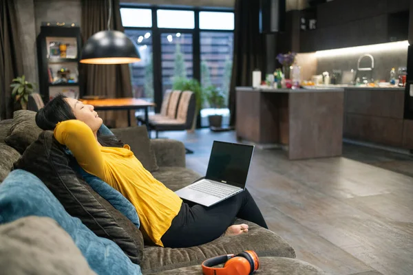 Oturma odasındaki kanepede dizüstü bilgisayarla oturan güzel, rahat Asyalı bir kadının görüntüsü.