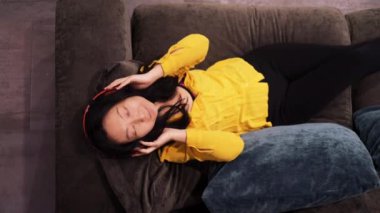 Asyalı genç bir kadın rahat koltukta, kulaklık takmış, gözleri kapalı rahat bir şekilde dinleniyor. Asyalı bir kız, evde rahat bir müzik ve müzik dinlemeyi seviyor. Üst görünüm yavaş çekimde.
