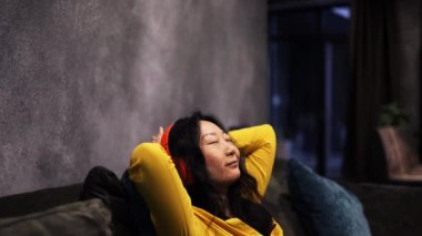 Genç Asyalı Kadın Gözleri Kapalı Evdeki Koltukta Uzanıyor ve Kulaklıkla Müzik Dinliyor. Kadın Dinleniyor ve İnternetten Radyoyu Dinliyor. Rahatlama, Meditasyon ve Farkındalık.