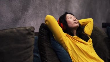 Genç Asyalı Kadın Gözleri Kapalı Evdeki Koltukta Uzanıyor ve Kulaklıkla Müzik Dinliyor. Kadın Dinleniyor ve İnternetten Radyoyu Dinliyor. Rahatlama, Meditasyon ve Farkındalık.
